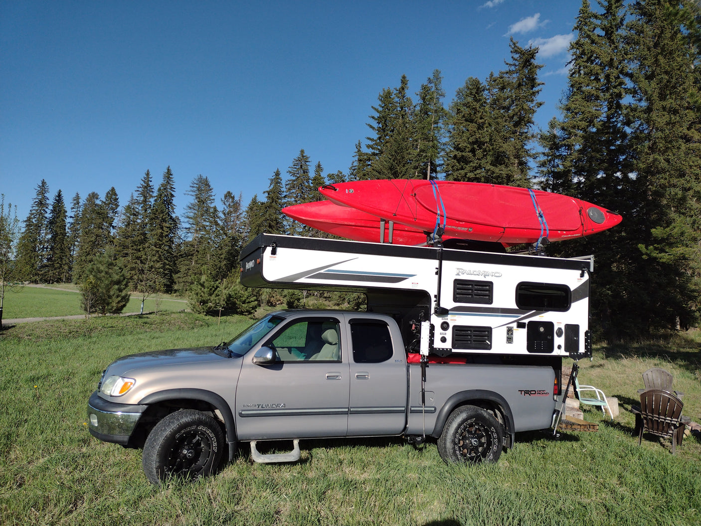 RV rental-ready Toyota Tundra truck with a mounted Palomino truck camper and two red kayaks, parked in a grassy field near pine trees.