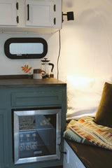 Inside a cozy RV rental: mini-fridge, coffee maker, and comfortable seating with patterned blanket and pillows.