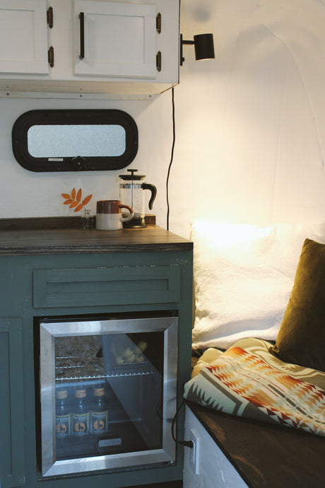 Inside a cozy RV rental: mini-fridge, coffee maker, and comfortable seating with patterned blanket and pillows.