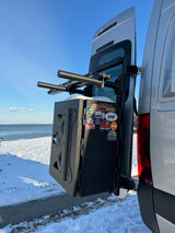 RV rental van's rear: cargo box with park stickers & ski rack visible. Parked on snowy ground by the ocean.