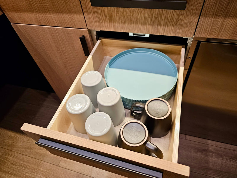 Open RV rental kitchen drawer with organized, compact storage of dishes: light cups, dark mugs, and a two-tone blue tray. — photo 12