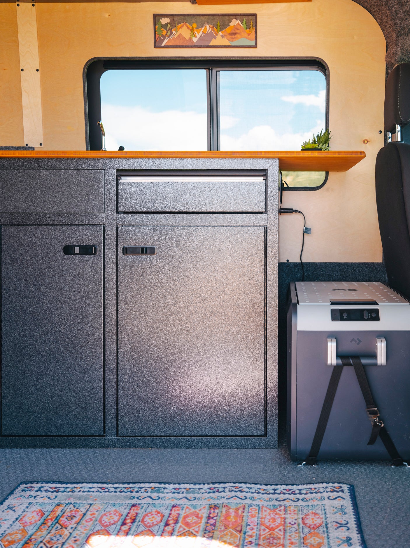 Cozy RV rental interior with textured cabinets, wood counters, a portable fridge, and a vibrant rug. Ideal for travels.