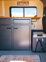 Cozy RV rental interior with textured cabinets, wood counters, a portable fridge, and a vibrant rug. Ideal for travels.