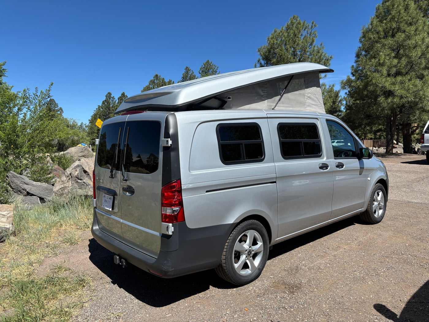 Silver pop-top Mercedes-Benz Metris camper van, an RV rental, parked on a sunny gravel road surrounded by pine trees. — photo 9