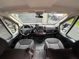 Inside an RV rental camper van: front cabin showing dashboard, steering wheel, seats, and outside view through the windshield.