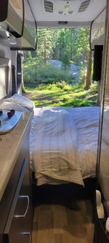Cozy rv rental interior with a bed looking out onto a stunning forest and stream view. Perfect for nature lovers.