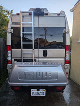 Rear of an RV rental van featuring a Thule cargo box, ladder, and California plate 9LDH234. Parked on a wet driveway.