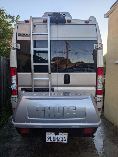Rear of an RV rental van featuring a Thule cargo box, ladder, and California plate 9LDH234. Parked on a wet driveway.