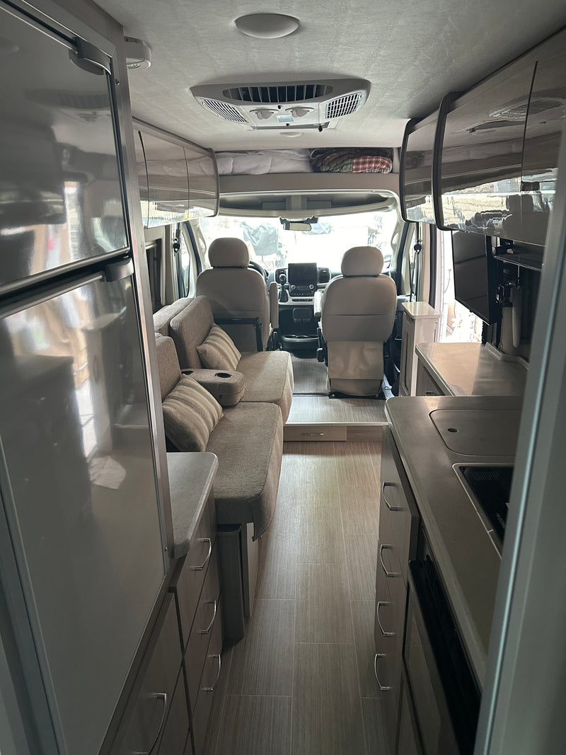 Bright RV rental interior, showing the living area with sofa, kitchen with counter, fridge, TV, and front cockpit. Overhead bunk visible. — photo 14