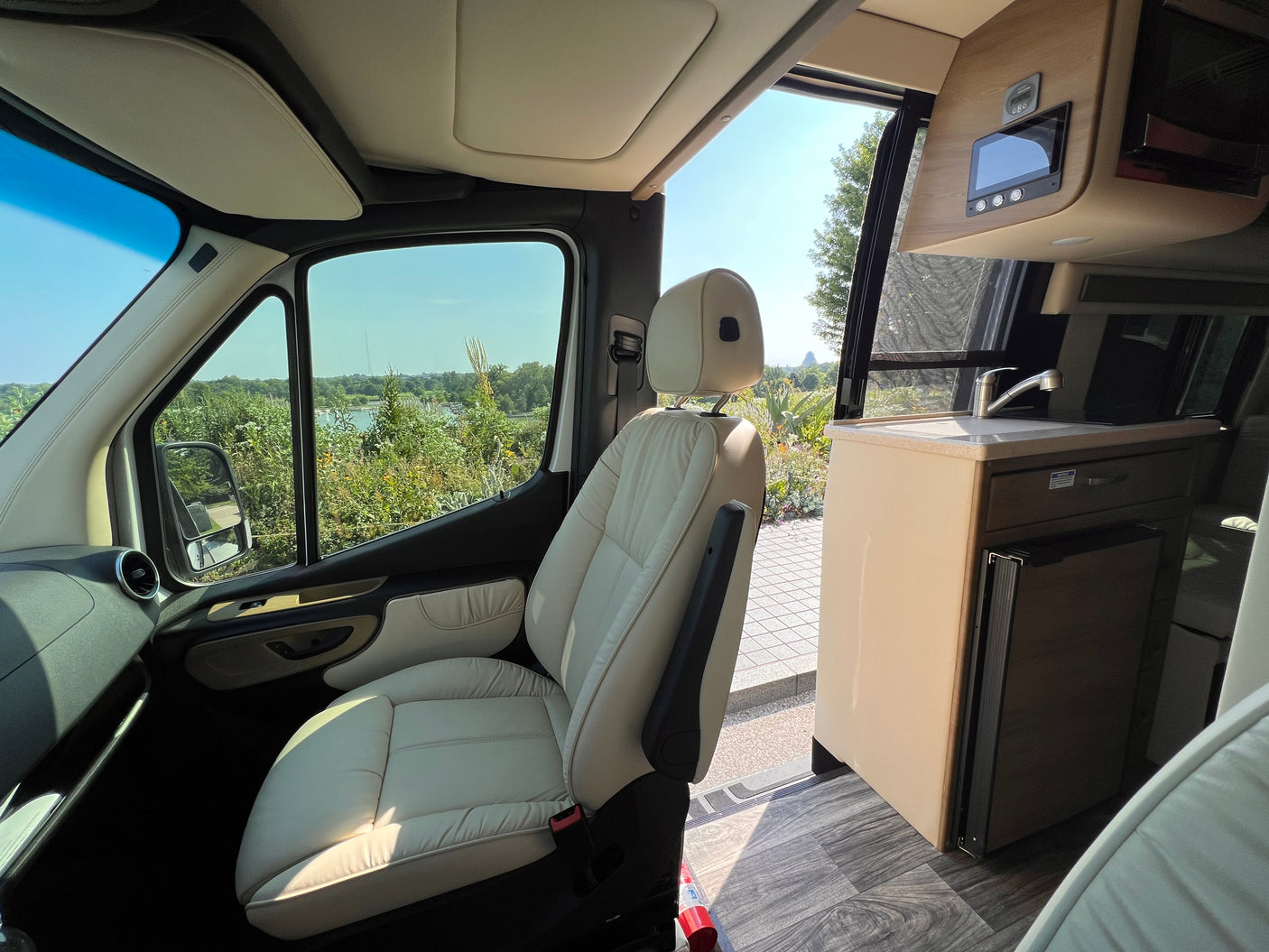 RV rental interior with a cream driver's seat, showcasing a scenic lake view, and a compact kitchen area.
