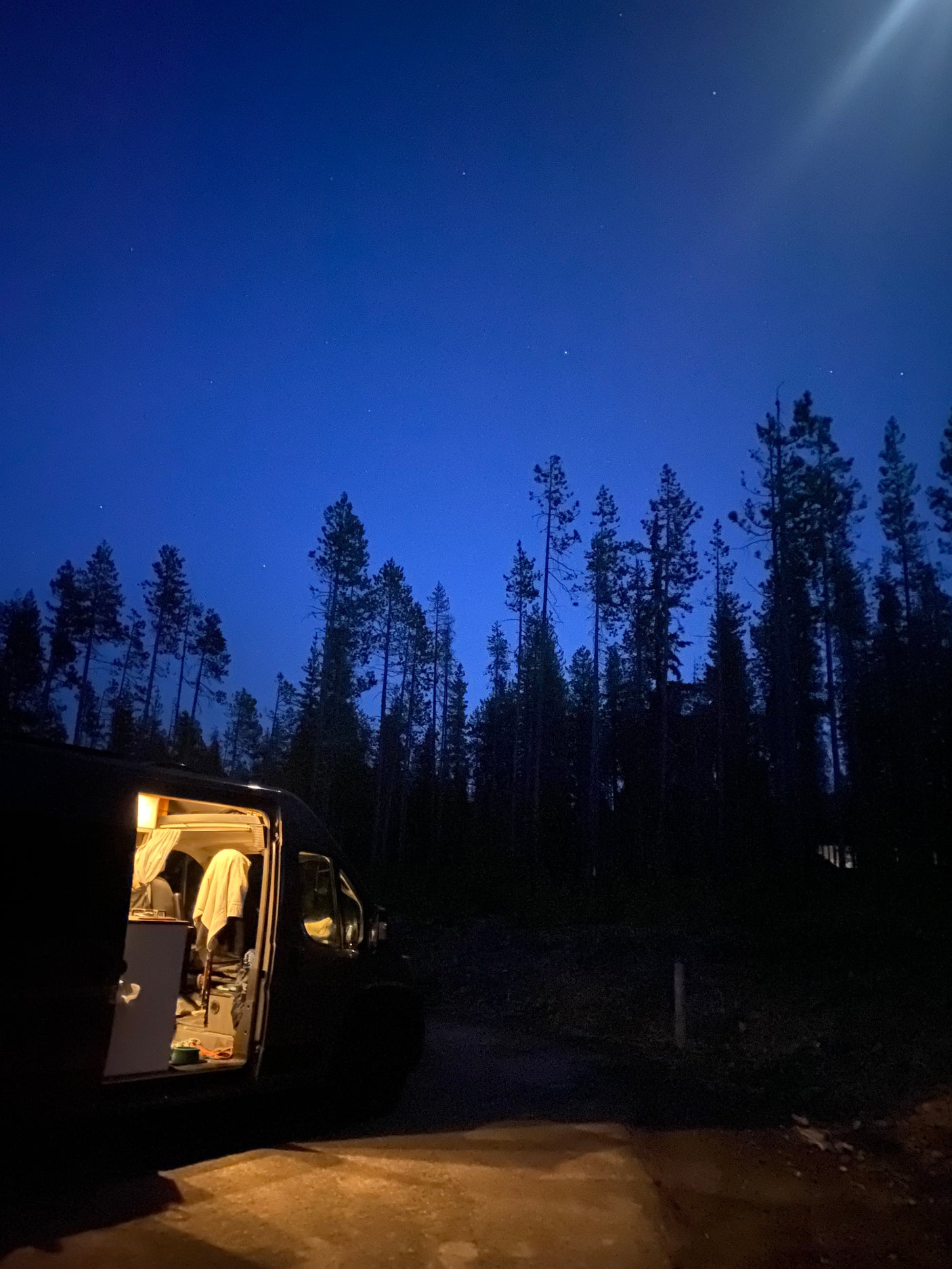 Night view: an RV rental with its interior lights glowing, nestled in a dark forest under a sky full of stars.