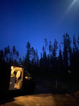 Night view: an RV rental with its interior lights glowing, nestled in a dark forest under a sky full of stars.