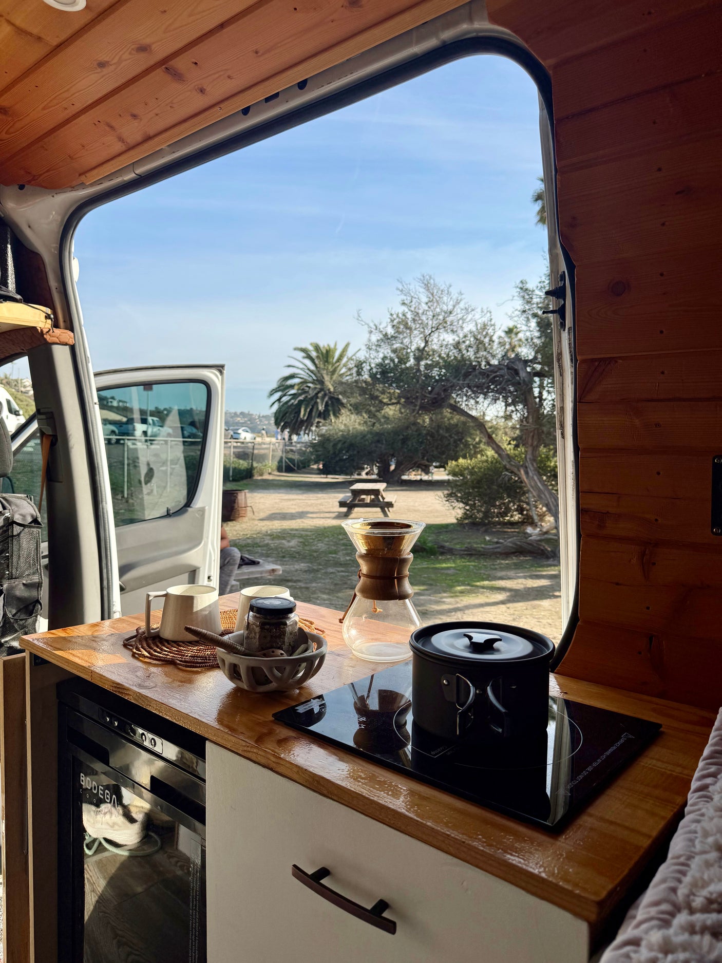 Morning coffee in an RV rental! A Chemex and pot are on the counter, with a vibrant view of trees, a picnic table, and blue sky.