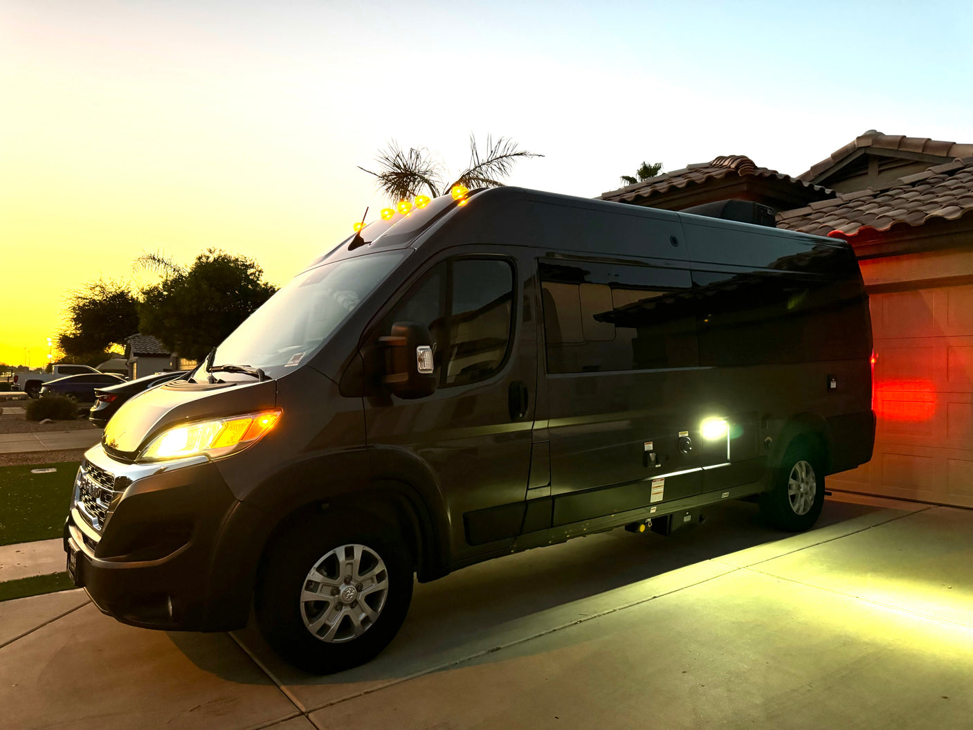 A dark grey camper van (RV rental) illuminates its driveway with bright side lights, amber roof lights, and headlights at dusk.