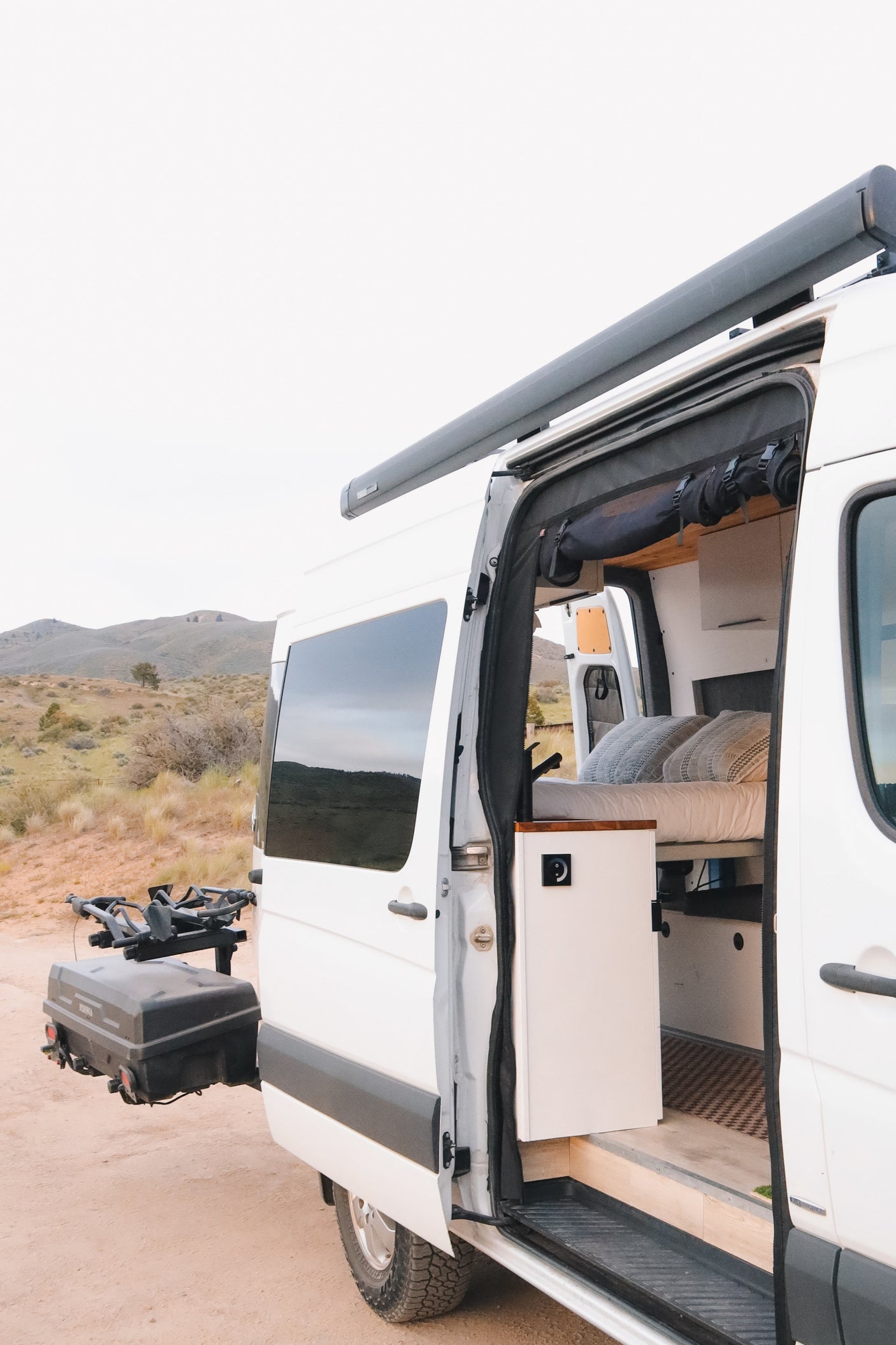This RV rental camper van has its side door open, revealing a bed, kitchen counter, and storage, parked in a dry landscape.