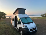 White Winnebago rv rental camper van with pop-top parked by a peaceful lake at sunrise. Side door open.