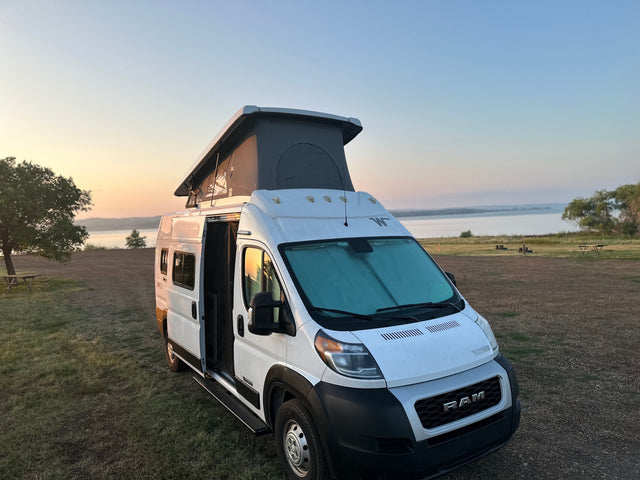 White Winnebago rv rental camper van with pop-top parked by a peaceful lake at sunrise. Side door open.