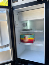 Inside an RV rental, an open mini-fridge reveals a 6-pack of beer and an ice tray, ready for a trip.