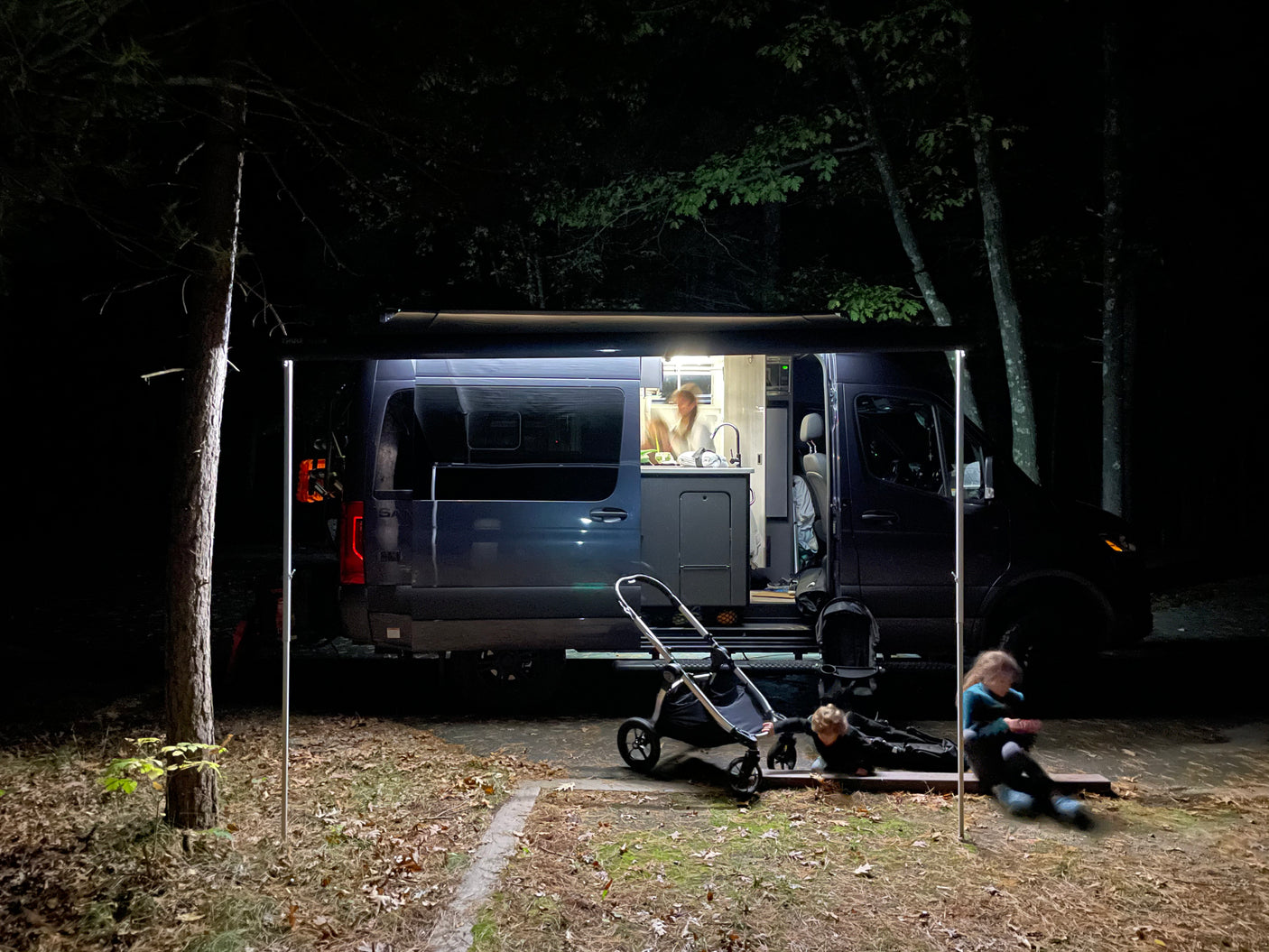 Nighttime fun with an RV rental: lit camper van with person at kitchen, and kids playing by stroller outside.