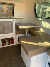 Bright RV rental interior showing a dinette with tan cushions, a small kitchen counter with fold-out table, and lower storage.