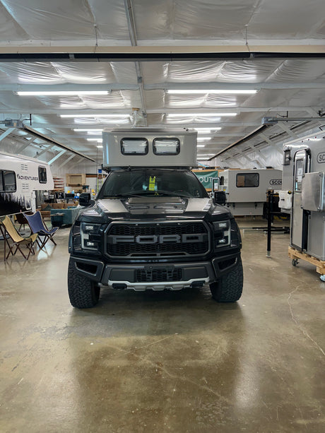 Black Ford Raptor truck with a light grey truck camper, available for rv rental, showcased in an RV manufacturing facility.