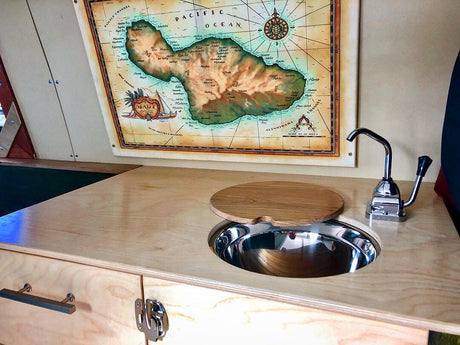 Compact RV rental kitchen features a sink with a wooden cover, hand pump faucet, and a large Maui map.