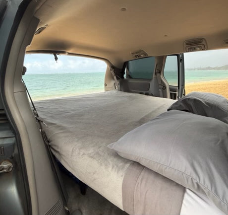 RV rental: Inside a customized van, a comfortable bed offers direct views of a sandy beach and blue ocean.