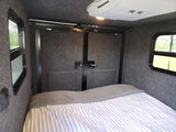 Cozy RV rental interior with bed, striped bedding, windows, storage nets, and overhead AC.