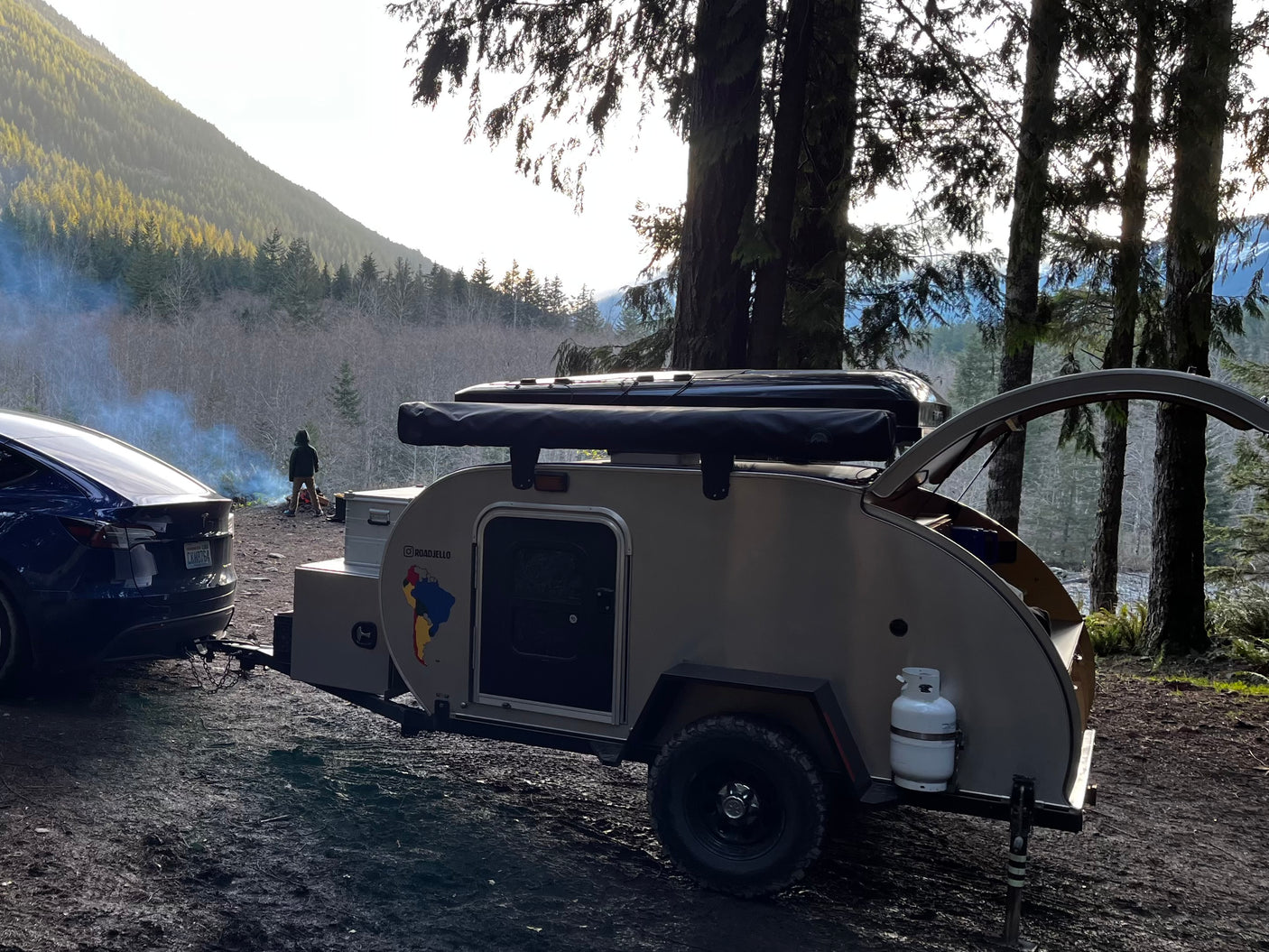 A dark blue Tesla tows a teardrop rv rental camper at a scenic forest campsite, with smoke rising from a distant campfire.