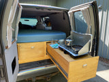 View inside an RV rental showing a full bed, custom wooden storage with a pull-out camping stove and kitchenware.