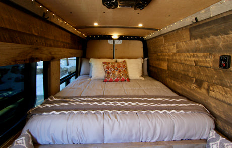 Rustic camper van bedroom for your rv rental. Features wood walls, warm string lights, comfy bedding, and a window view.