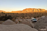 Enjoy an RV rental in the stunning Joshua Tree desert. White camper van near sunlit mountains, rocks, and Joshua trees.
