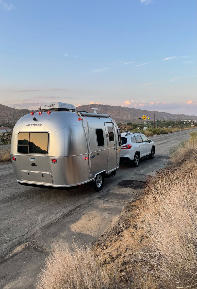  complete with an outdoor lounge setup under Joshua trees.|RV rental adventure: White SUV towing a sleek Airstream travel trailer at golden hour in a vast desert landscape.|White Subaru SUV towing an Airstream travel trailer — photo 2
