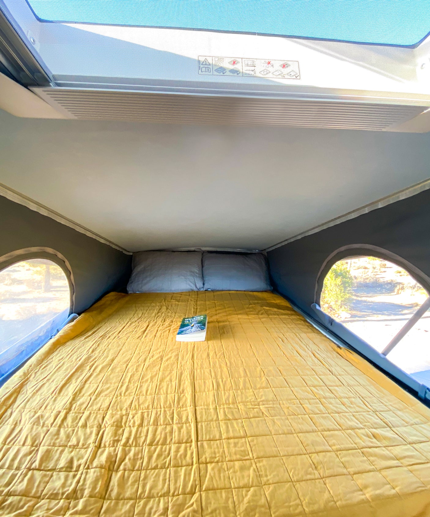 Inside an RV rental: a cozy yellow quilted bed with pillows and a book, framed by scenic side windows.