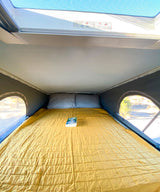 Inside an RV rental: a cozy yellow quilted bed with pillows and a book, framed by scenic side windows.