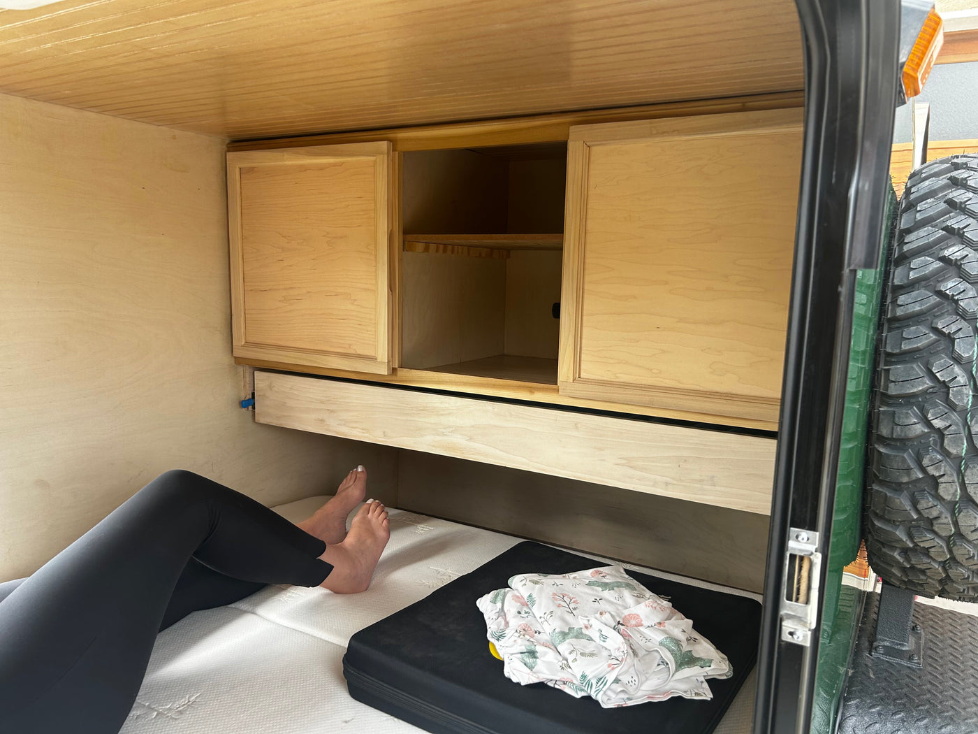 Cozy rv rental camper interior: person relaxing on a bed next to wooden cabinetry, shelves, and a storage drawer. — photo 6