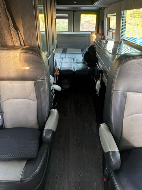 Inside an RV rental: front seats, dark wood floor leading to a kitchenette and convertible seating/bed area. Ideal for travel.