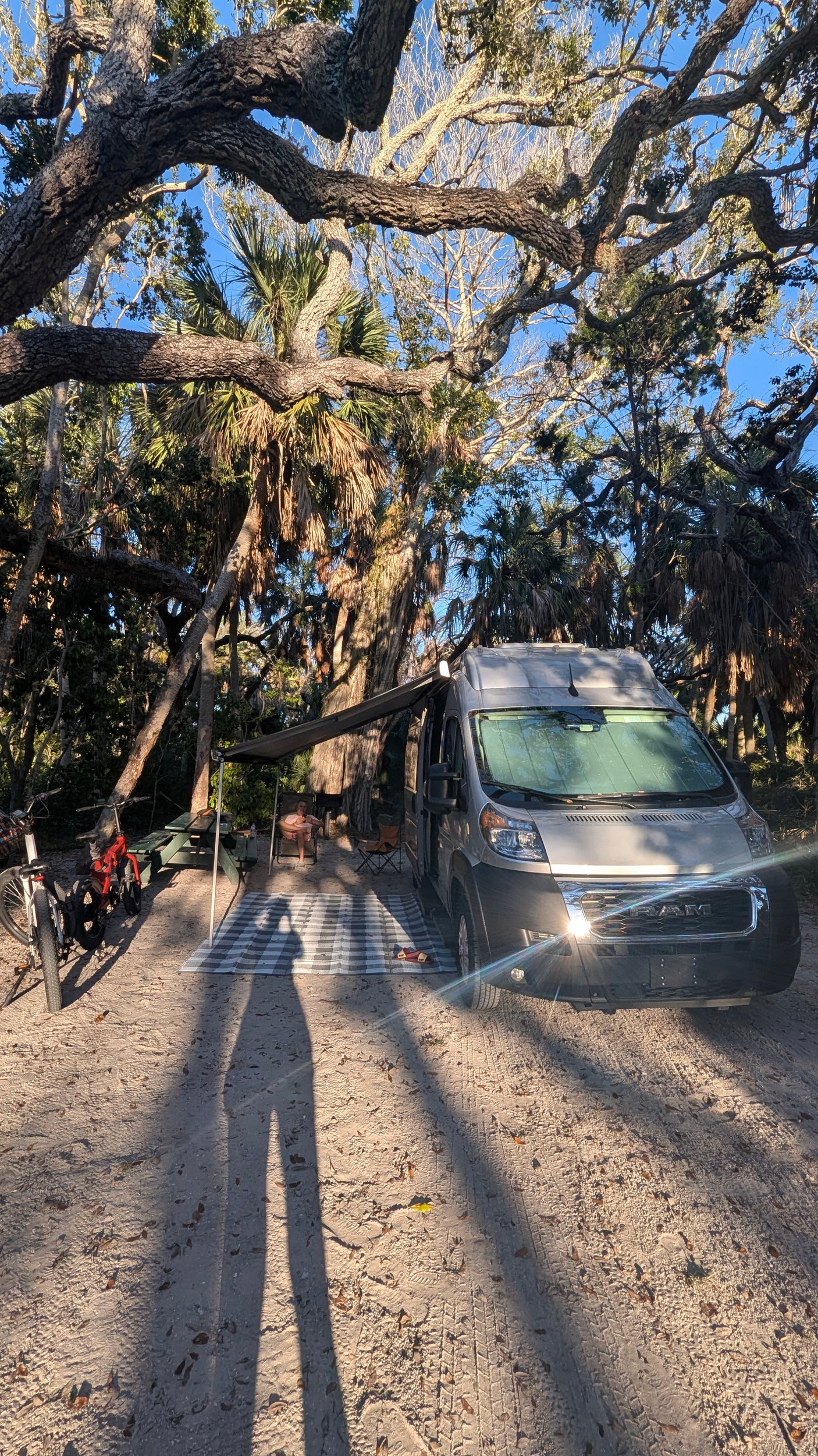 RV rental campervan at a sunny campsite with an extended awning, picnic setup, and bikes under large trees.