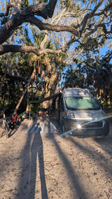 RV rental campervan at a sunny campsite with an extended awning, picnic setup, and bikes under large trees.