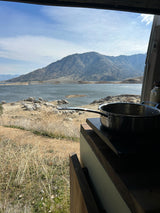 RV rental view: cooking with a lake & mountains backdrop. Enjoy van life with scenic outdoor cooking from your camper.