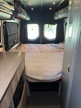 Cozy RV rental camper van bedroom. Features a made bed, overhead storage, and view out the rear doors.