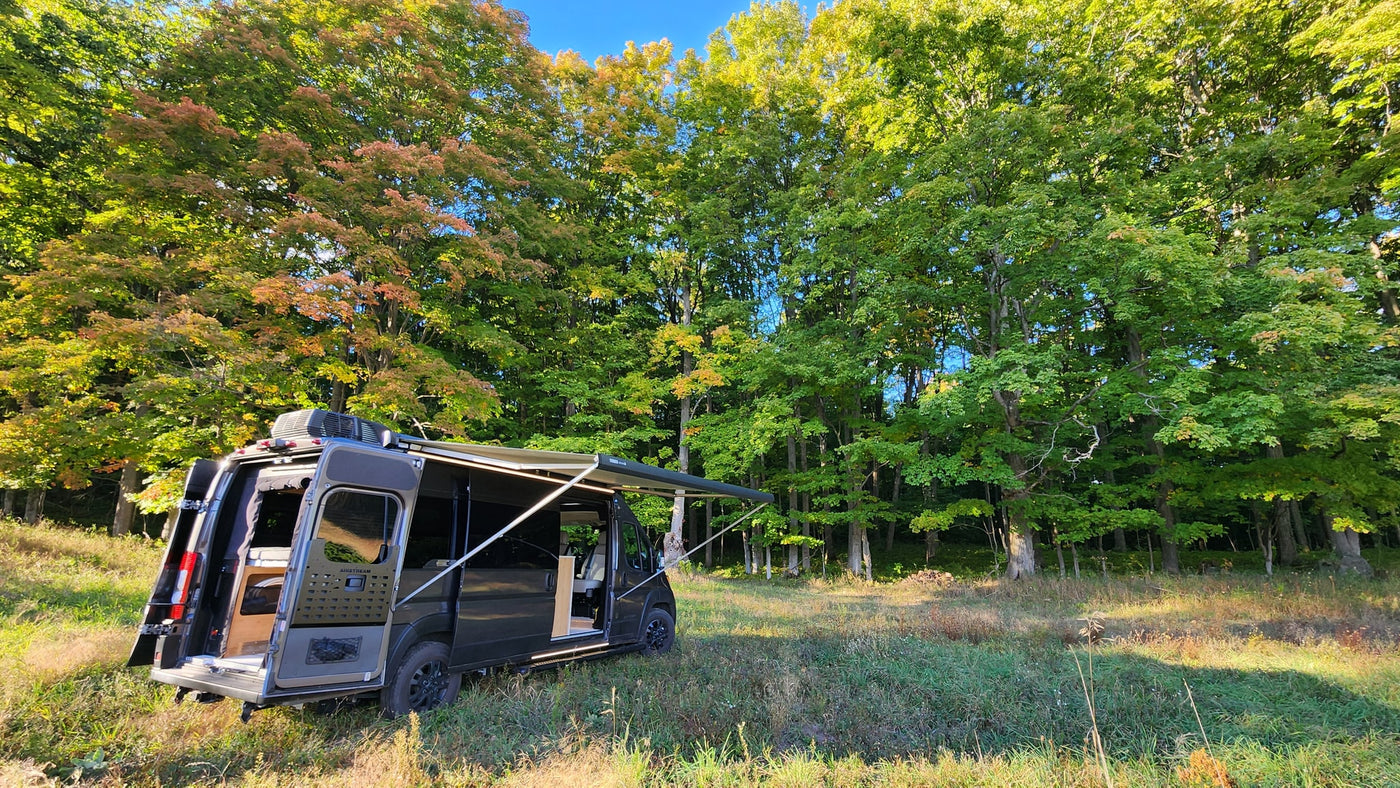 RV rental adventure: Dark camper van parked in a sunny, grassy clearing with open doors & extended awning, surrounded by fall trees. — photo 14