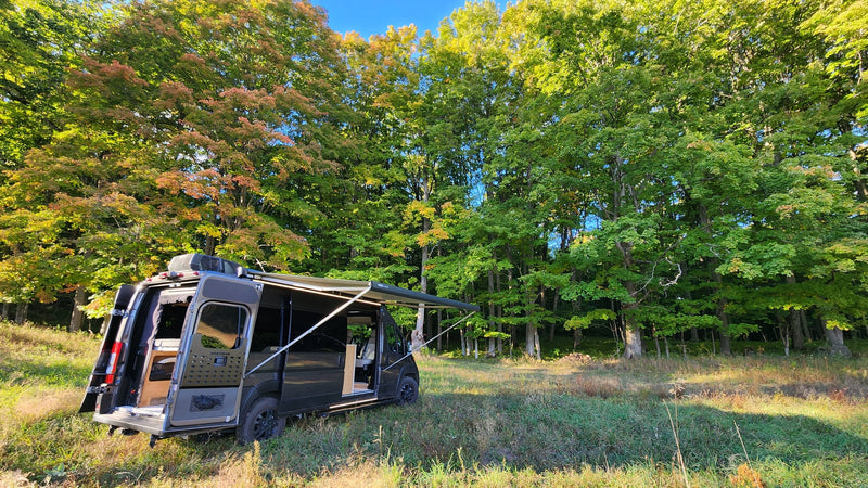 RV rental adventure: Dark camper van parked in a sunny, grassy clearing with open doors & extended awning, surrounded by fall trees. — photo 14