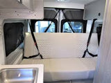 Inside an RV rental camper van, showing a white rear bench seat with seatbelts & a small sink area.
