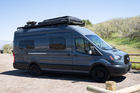 Dark gray camper van with roof rack, grill, and tent, ideal for an RV rental outdoor adventure.