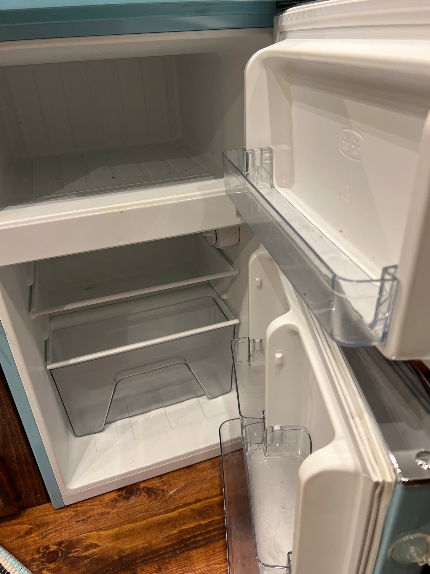 Clean, empty compact RV rental refrigerator. Inside features a freezer, clear shelves, and crisper drawer.