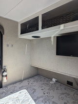 Cozy RV rental bedroom features a patterned mattress, overhead netted storage, wall-mounted TV, and fire extinguisher.