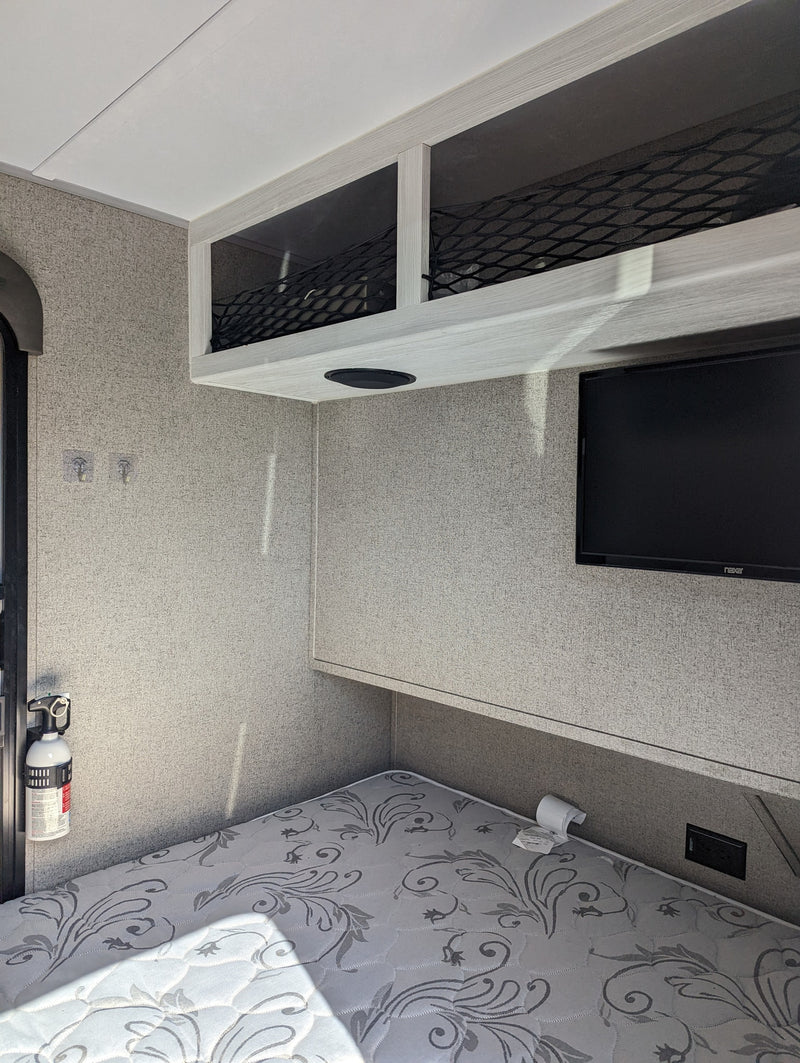 Cozy RV rental bedroom features a patterned mattress, overhead netted storage, wall-mounted TV, and fire extinguisher. — photo 11