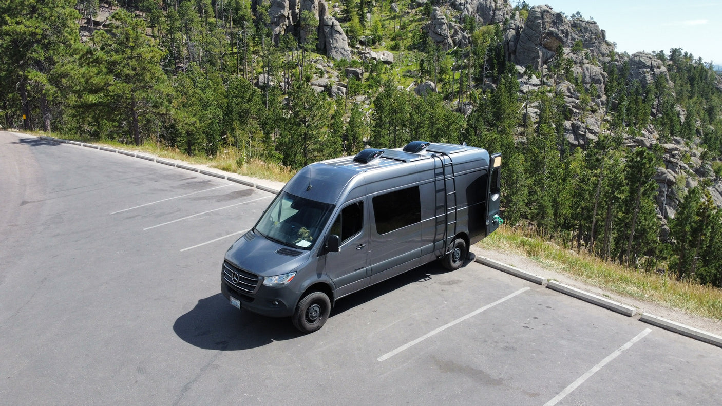 An RV rental camper van parked in a mountain lot, surrounded by a lush forest and rocky formations. Ideal for nature travel.