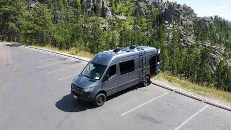 An RV rental camper van parked in a mountain lot, surrounded by a lush forest and rocky formations. Ideal for nature travel.